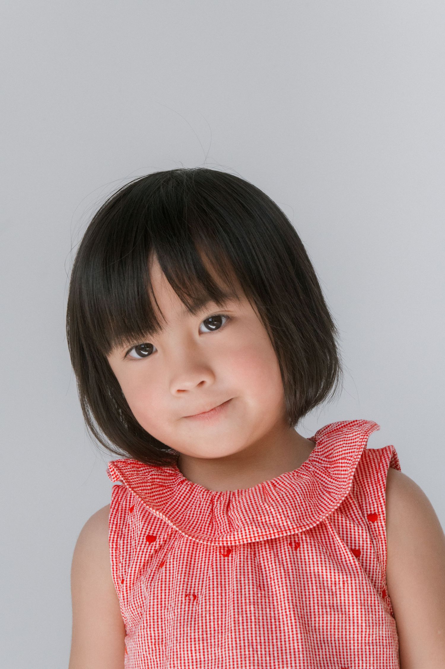 A young Asian girl with a bob haircut wearing a red gingham ruffle dress smiles softly against a gray background.