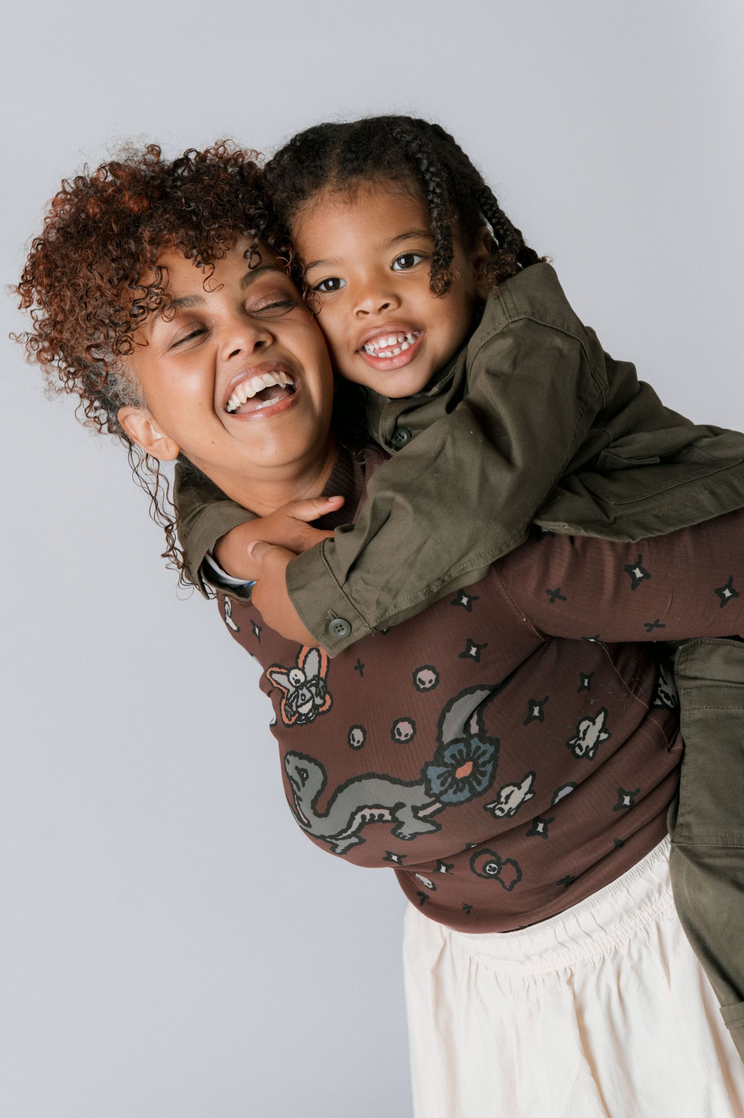 A joyful mother gives her smiling young daughter a piggyback ride against a light gray background.