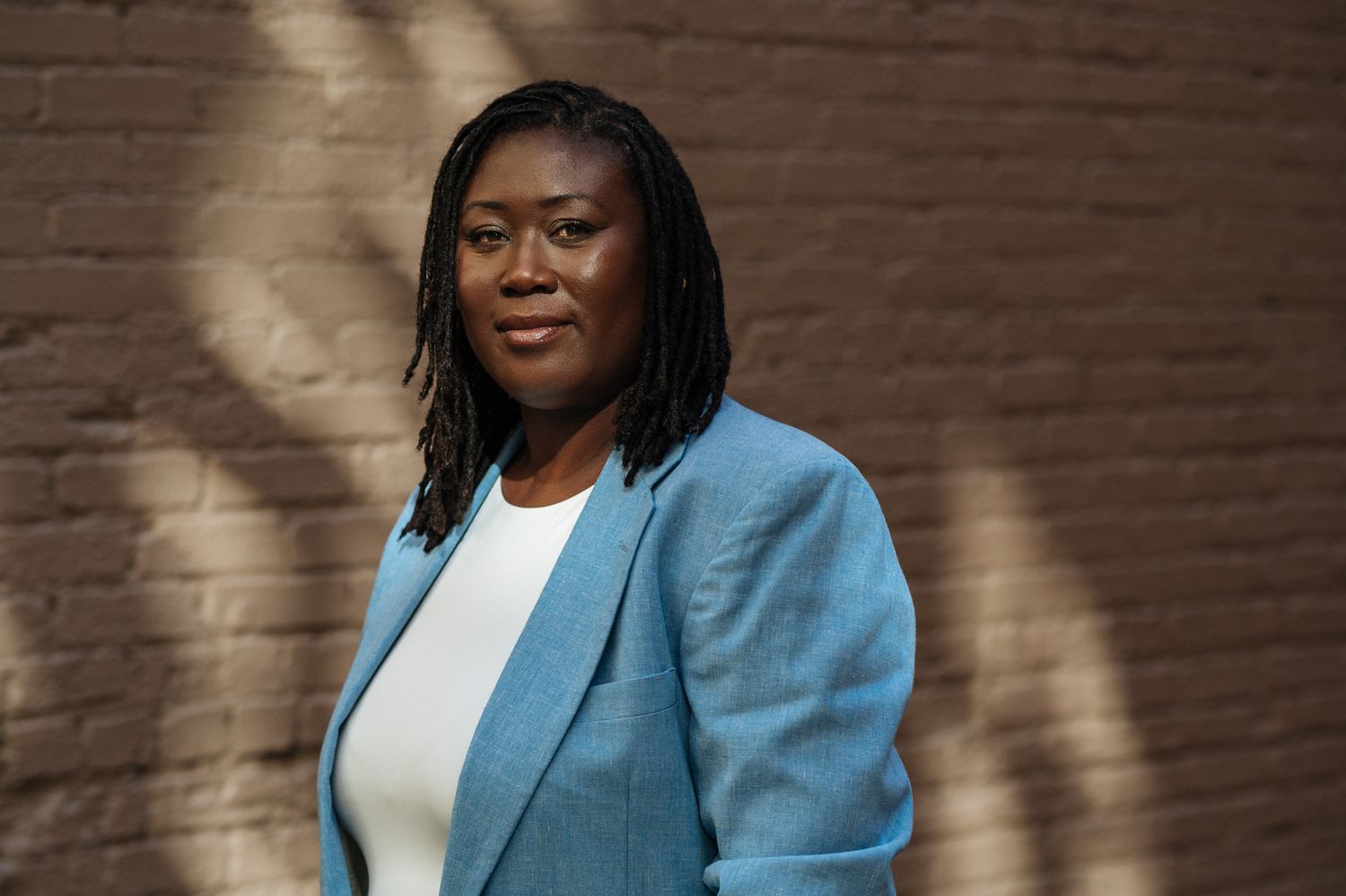 Professional woman in blue blazer posing confidently against a brick wall in natural sunlight.