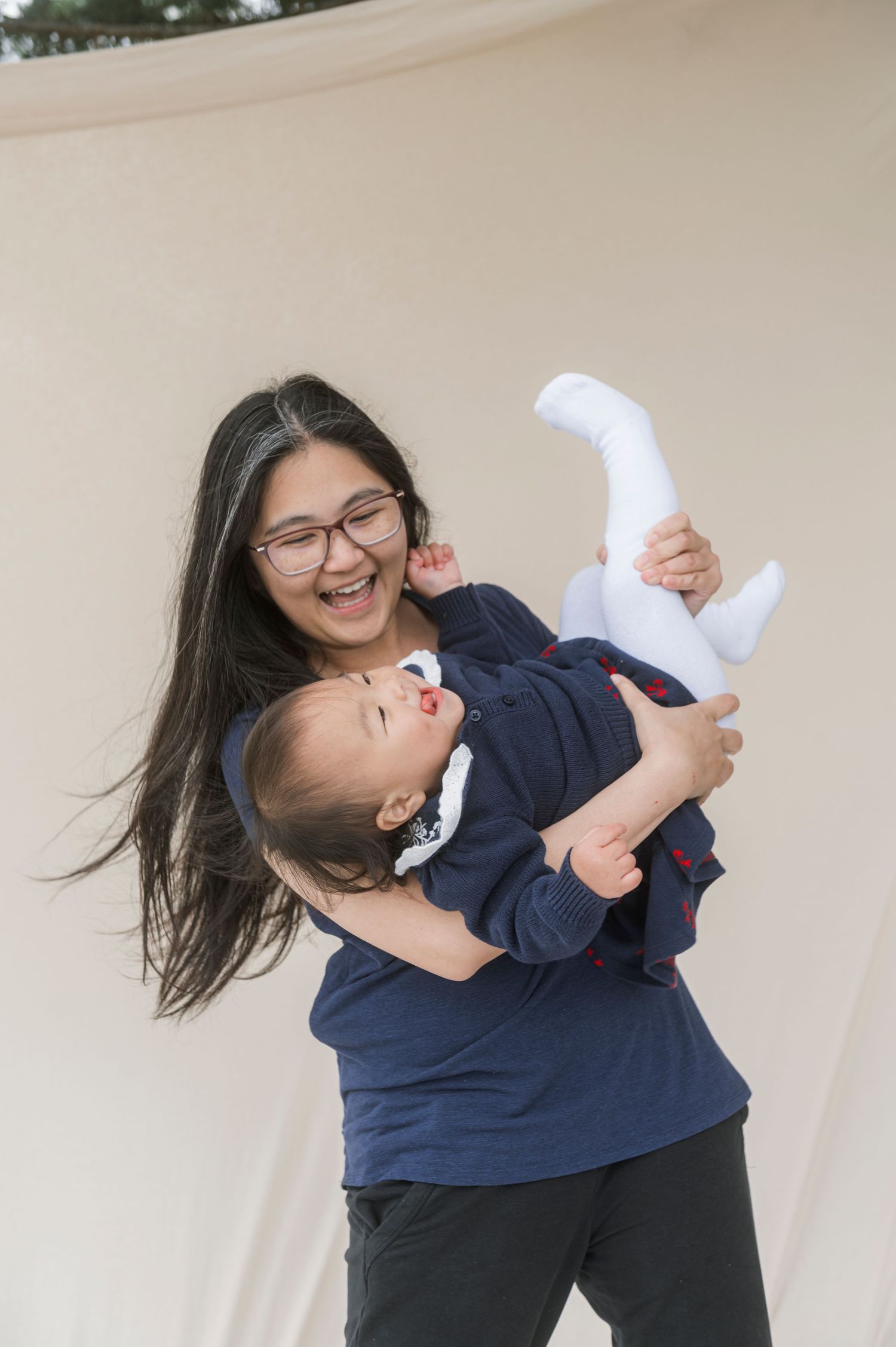 Smiling Asian woman with glasses joyfully holding a laughing baby in a navy outfit against a beige backdrop.