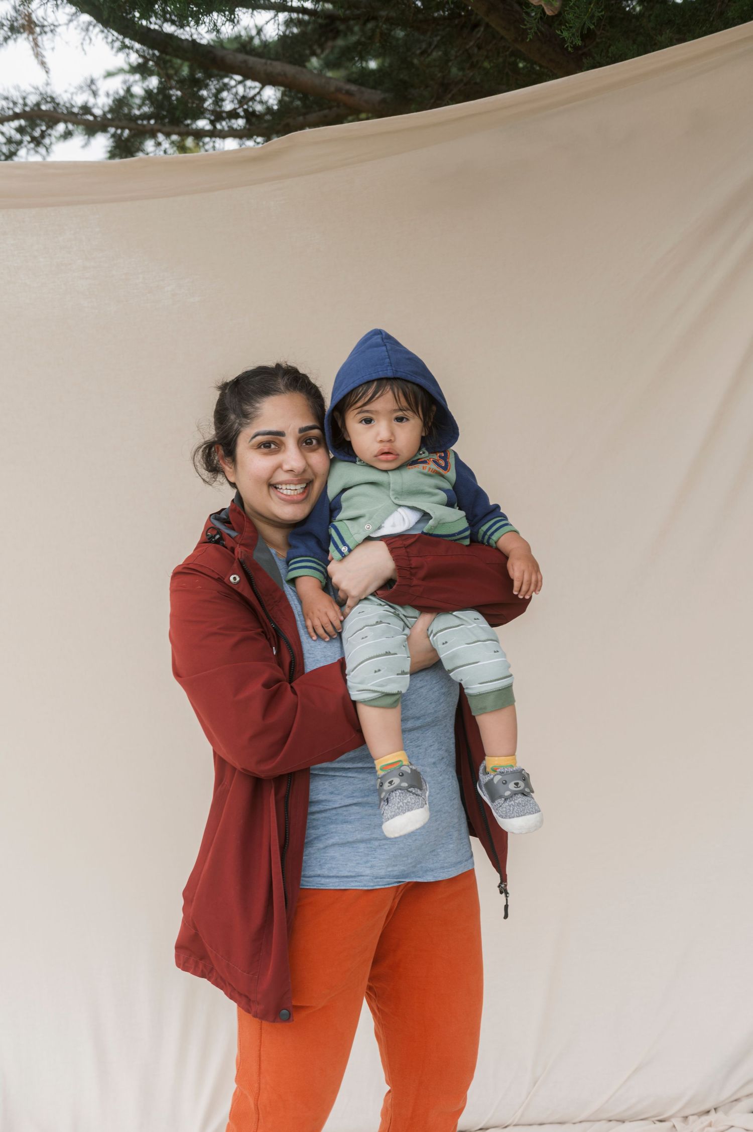 A smiling woman in a red jacket holds a baby in a blue hoodie outdoors near a canvas backdrop.