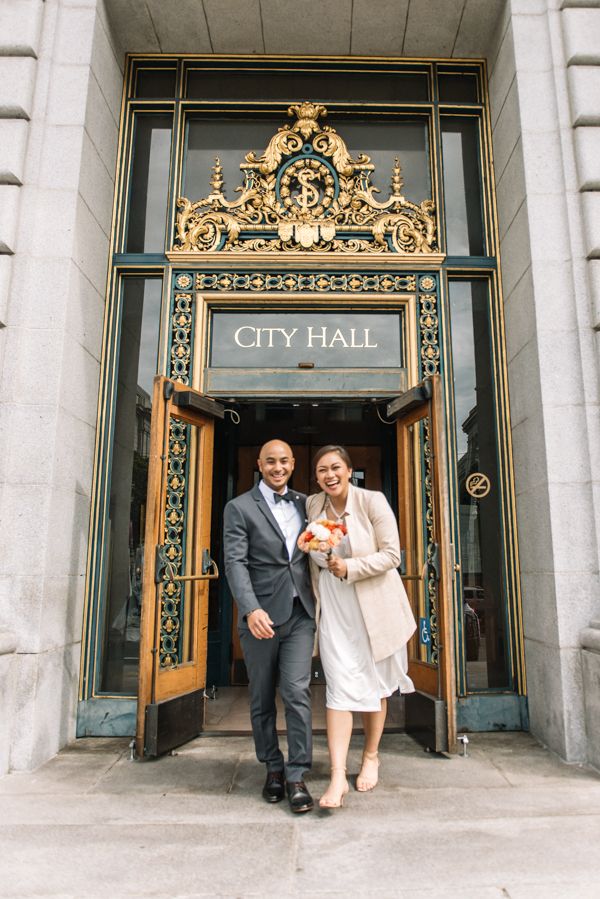 A couple poses in formal attire at the ornate gold-decorated entrance of City Hall for a wedding photo.