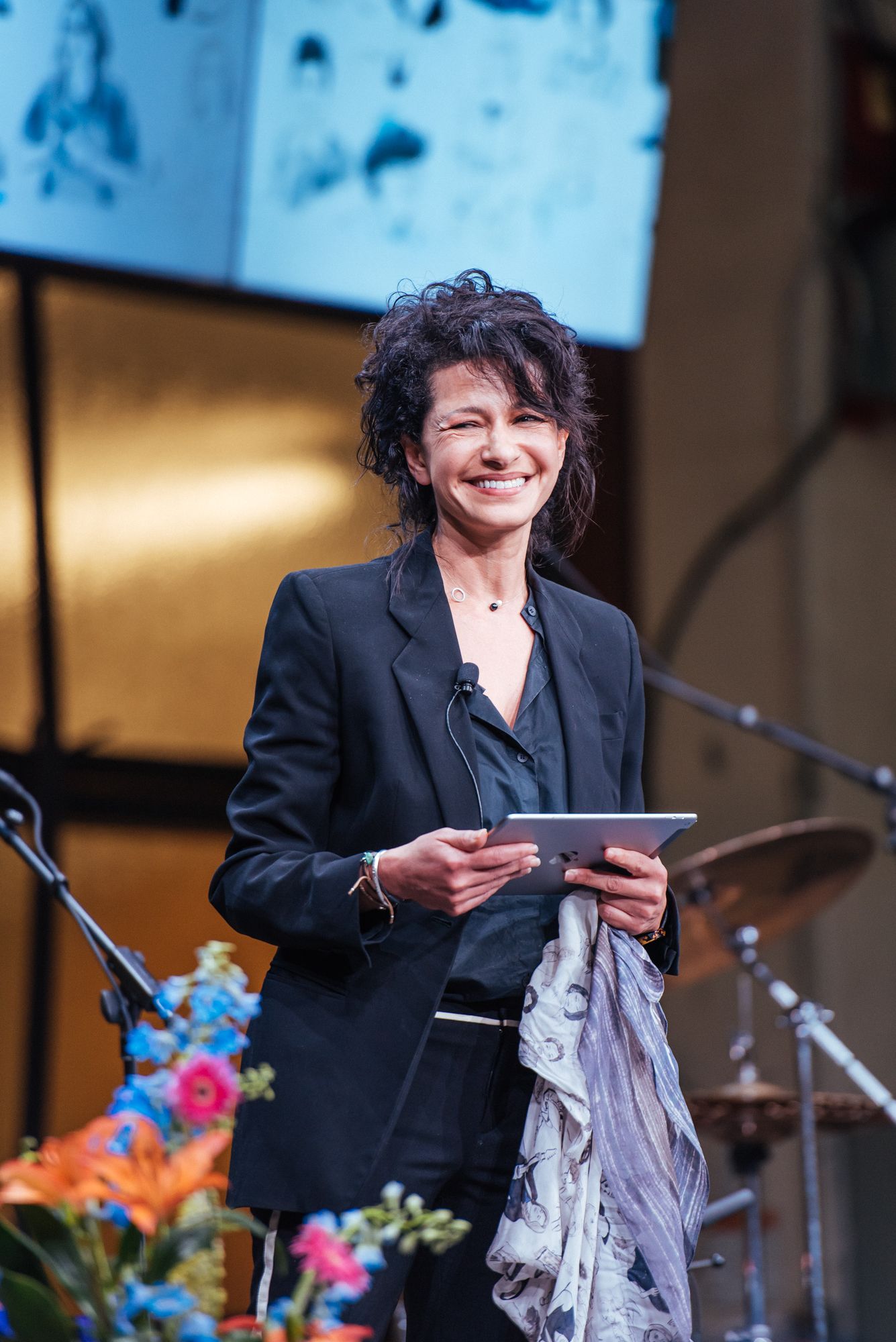 Person in black blazer speaking at podium with colorful flowers on stage during performance.