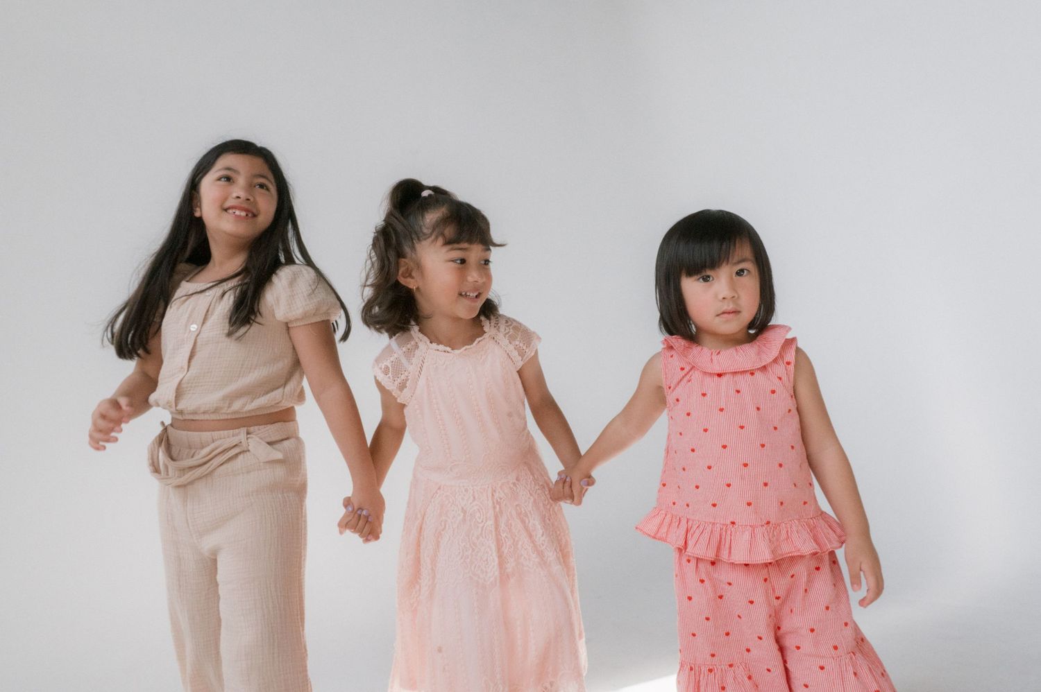 Three young Asian girls in pink and beige outfits hold hands and smile against a white background.