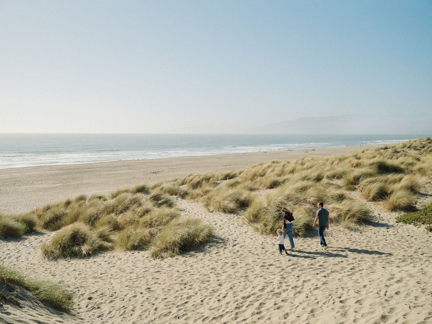 Two people walking on sandy beach dunes with ocean waves and clear sky in the background.