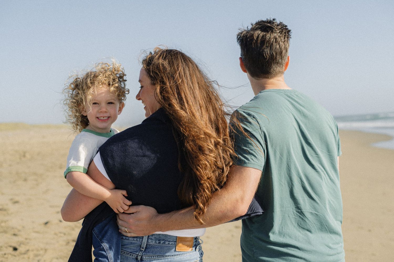 Family of three enjoying a sunny beach day, with parents embracing while holding their curly-haired child.