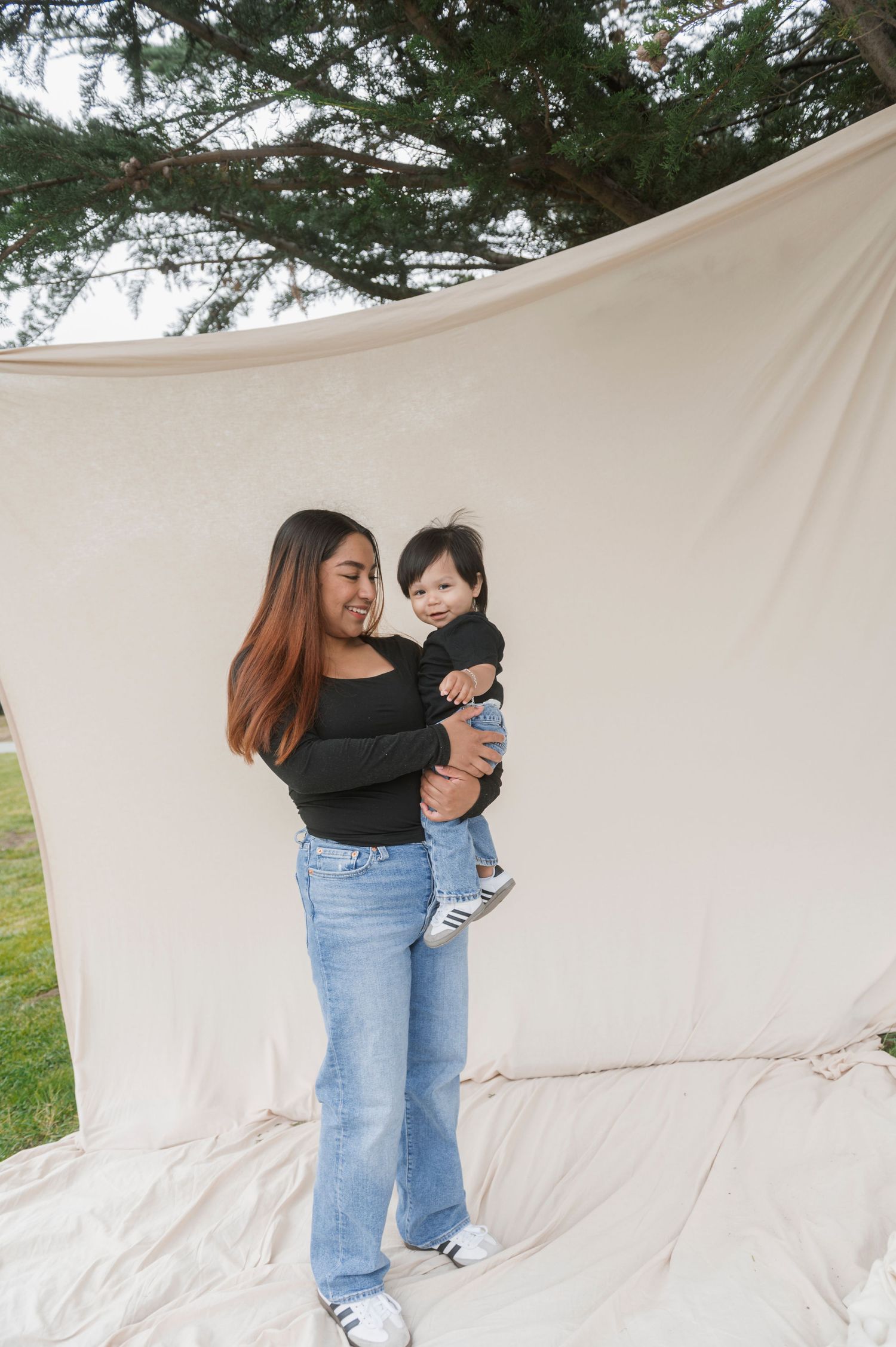 A young woman in black top and jeans holds a smiling baby outdoors against a white fabric backdrop.