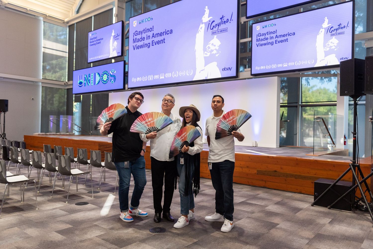 Four people pose with colorful fans at a Made in America Viewing Event in a modern venue with large screens.