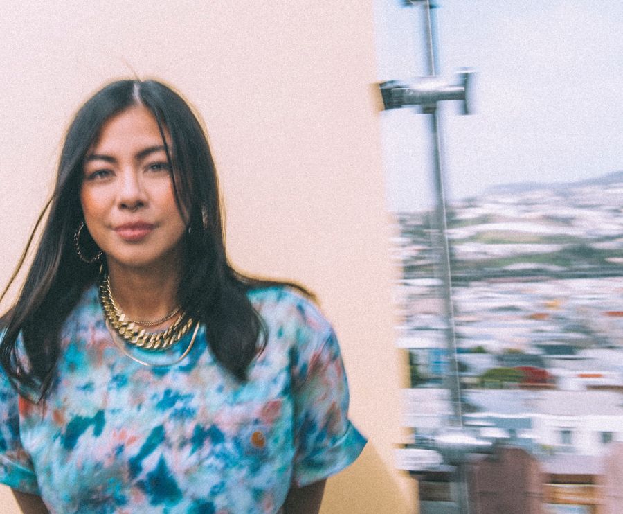 Woman in floral blue top and gold necklace posing outdoors with a blurred cityscape in the background.