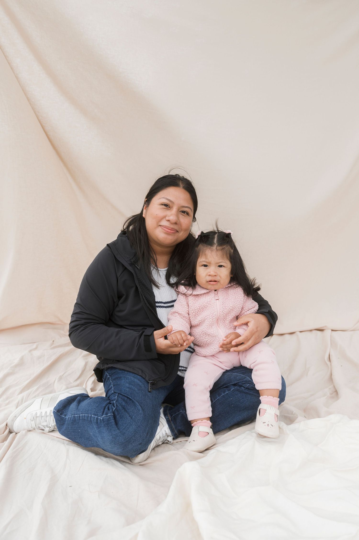 A woman in a black jacket sits with a toddler in a pink outfit on white fabric before a cream backdrop.