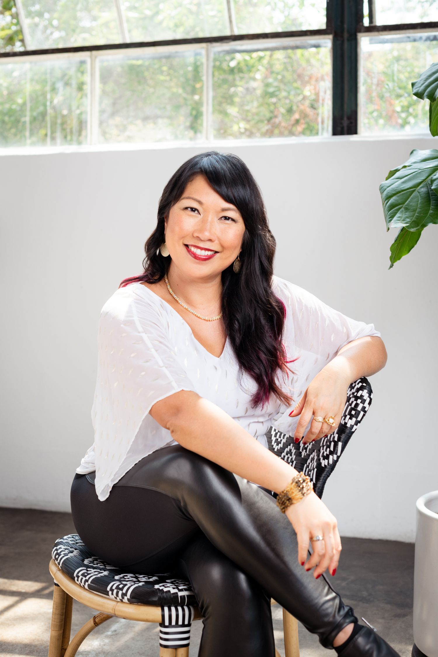 Smiling Asian woman in white top and black leather pants sitting on a stool in a bright modern studio.