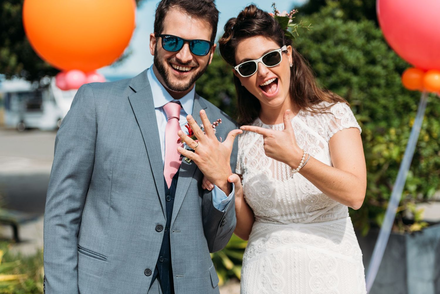 A happy couple in wedding attire holds colorful balloons outdoors on a sunny day.