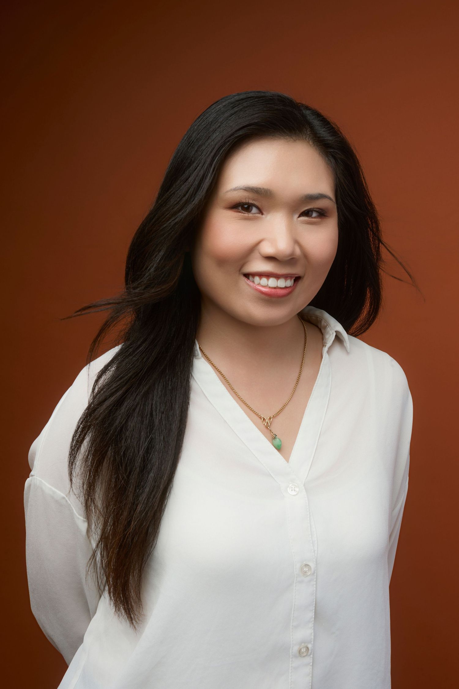 Smiling Asian woman with long black hair wearing a white blouse and gold necklace against a brown background.