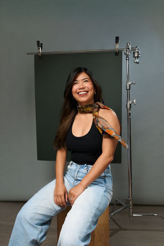 Smiling woman in black tank top, jeans, and printed scarf sits in photography studio with green backdrop.