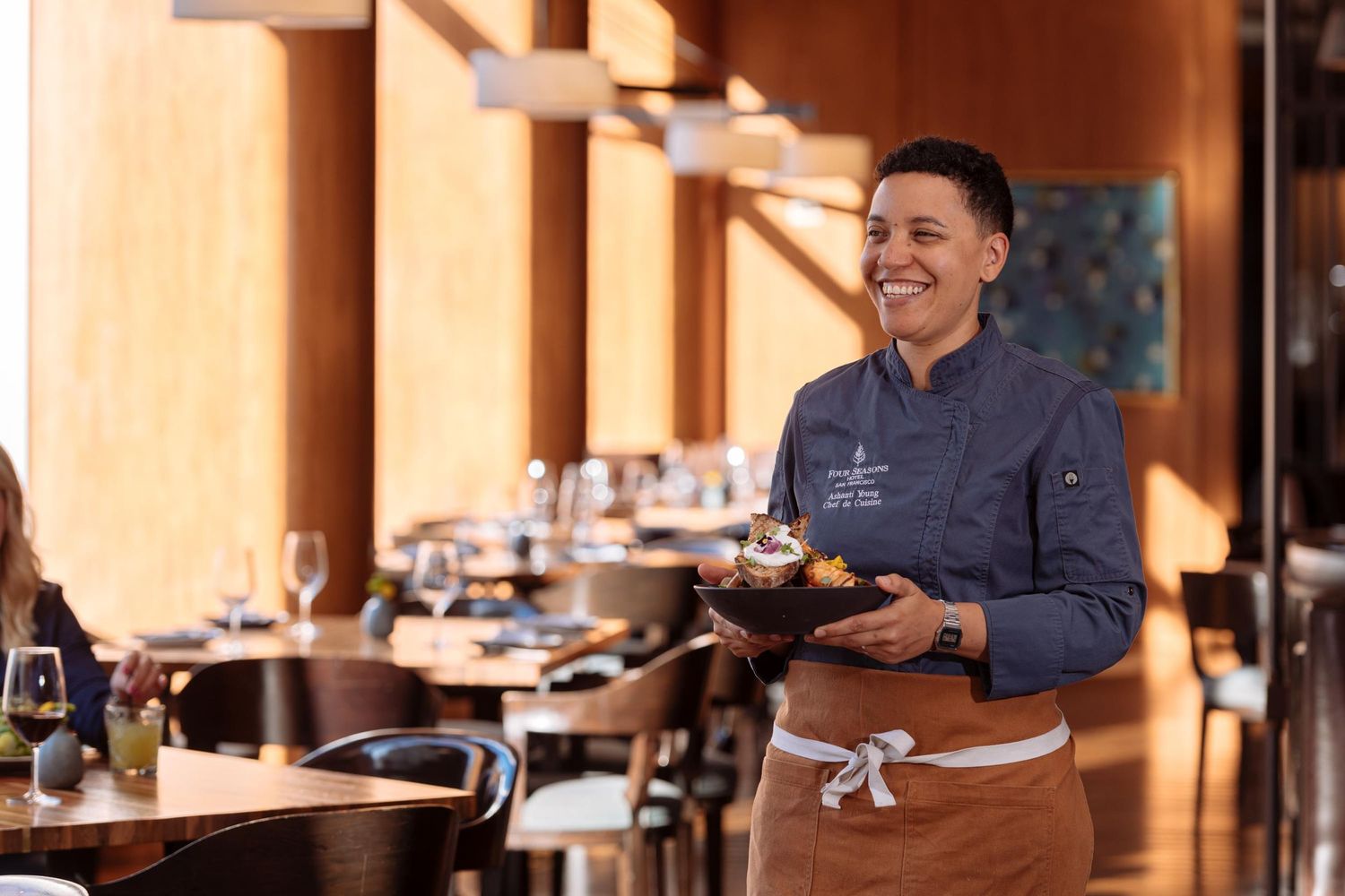 Smiling chef in blue uniform and brown apron carries a dish through a modern restaurant dining room.