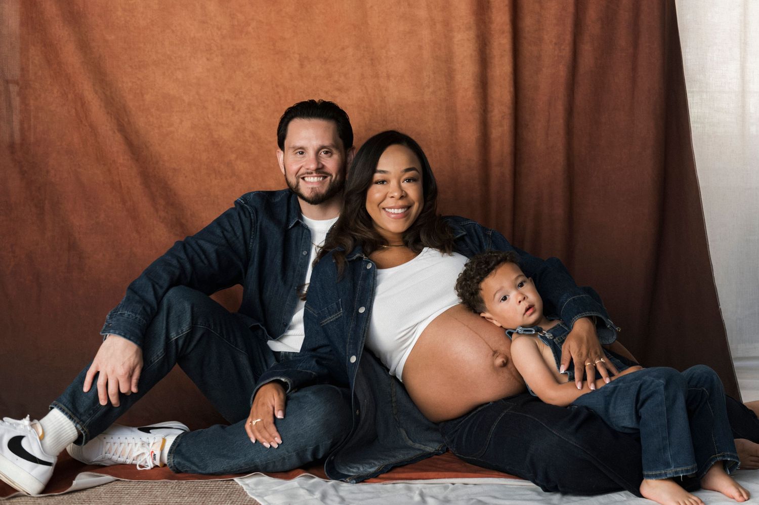 Expecting family maternity photo with parents and toddler posing against a warm brown backdrop.