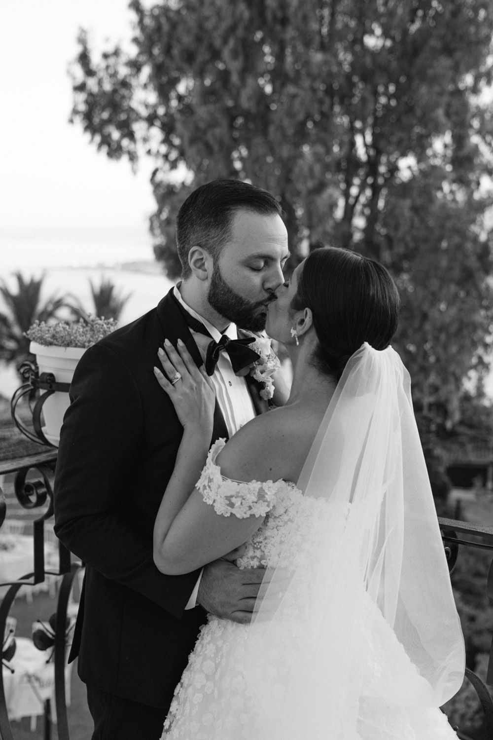 Black and white photo of a bride and groom sharing a romantic kiss on a balcony surrounded by trees in Taormina at Villa Mon Repos.