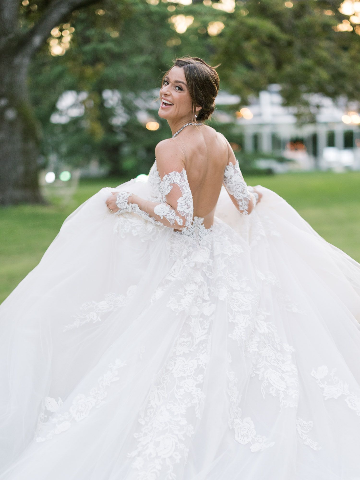 A bride in a stunning backless lace wedding gown with long sleeves twirls joyfully in a garden setting at sunset. Villa Cavazza.
