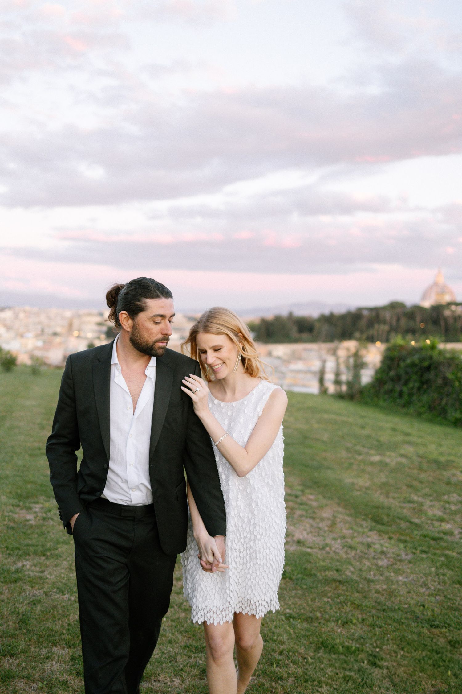 Romantic couple in formal attire walking together on grassy hillside during sunset. A surprise proposal in Italy.