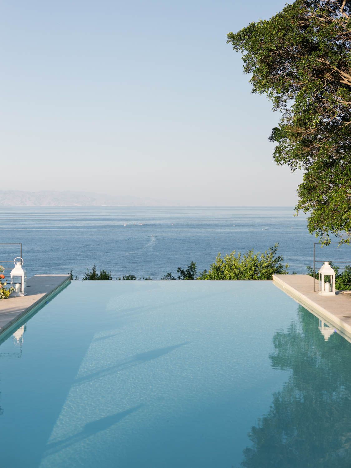 Stunning infinity pool overlooking a calm blue sea with lush trees and white lanterns on a clear day. Wedding at Villa Mon Repos, Taormina.