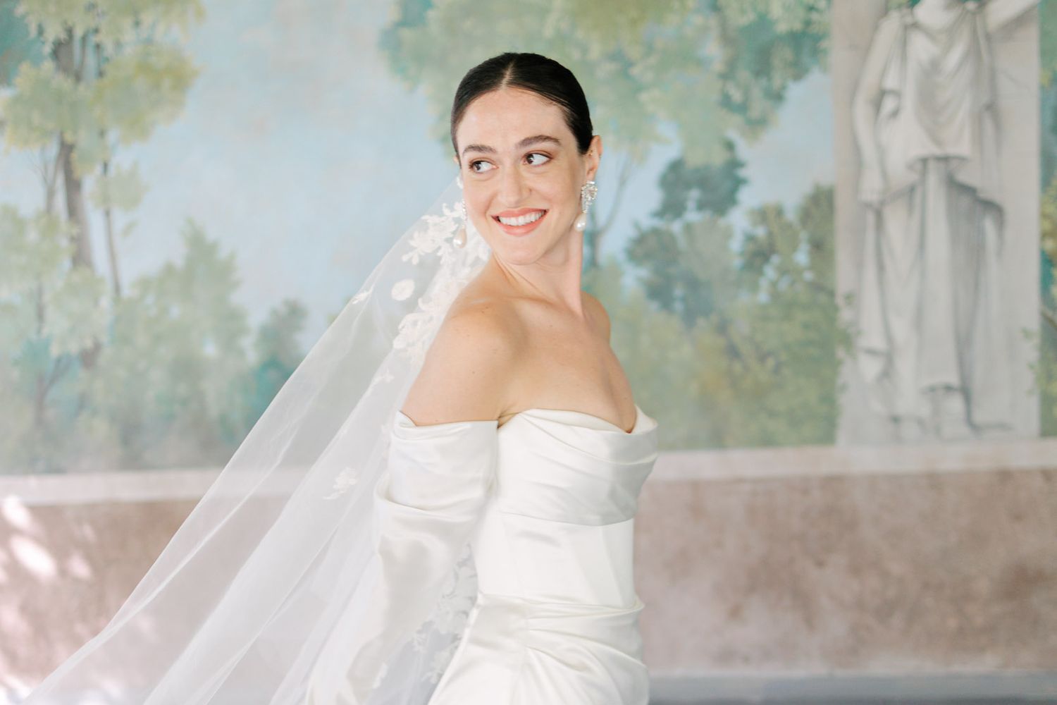 A bride in a strapless white wedding gown smiles while wearing a flowing chapel-length veil. Wedding in Rome