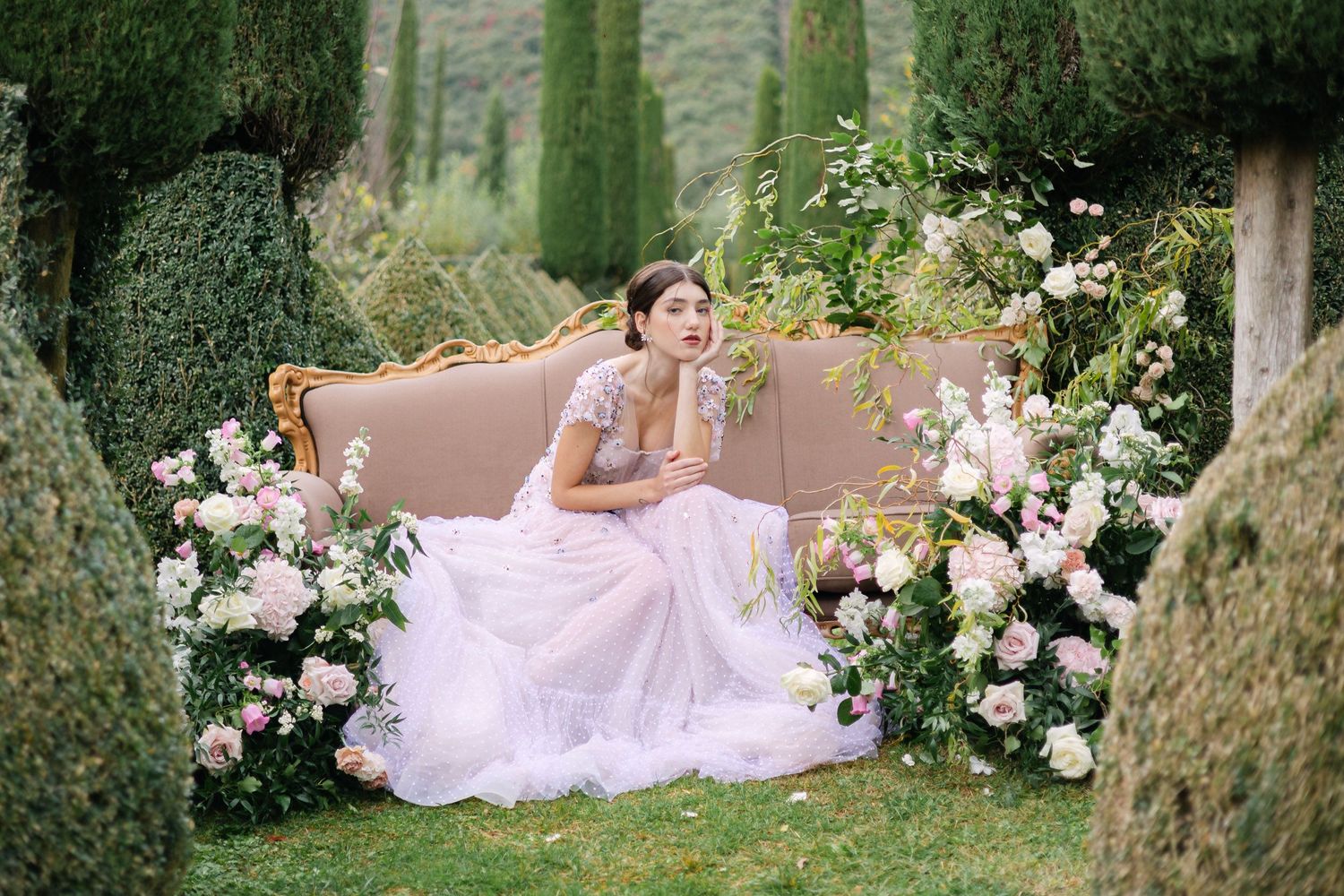 A person in a flowing white gown reclines gracefully among pink roses and greenery in an enchanted garden setting.