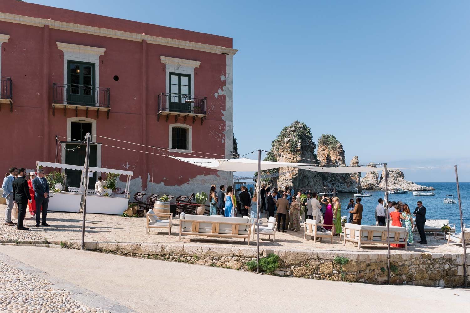 Outdoor wedding reception by the sea at Tonnara di Scopello in Sicily, Italy, with guests mingling on a sunny day.