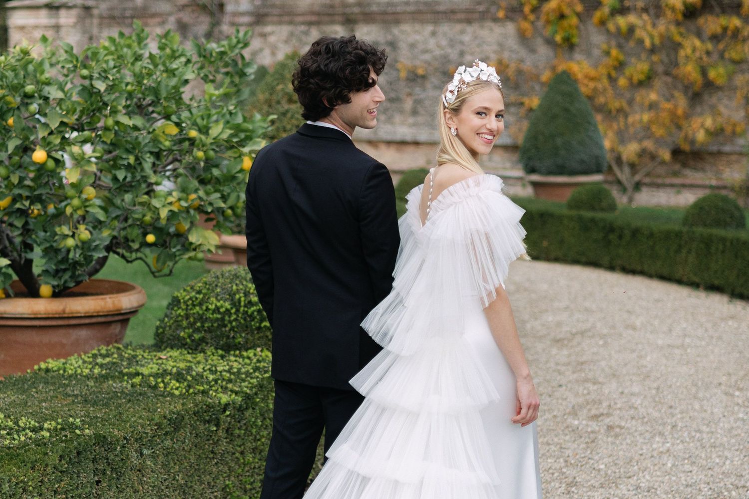 A couple in formal wedding attire standing on a gravel path in a manicured garden with greenery and stone walls. Villa Cetinale, Tuscany.