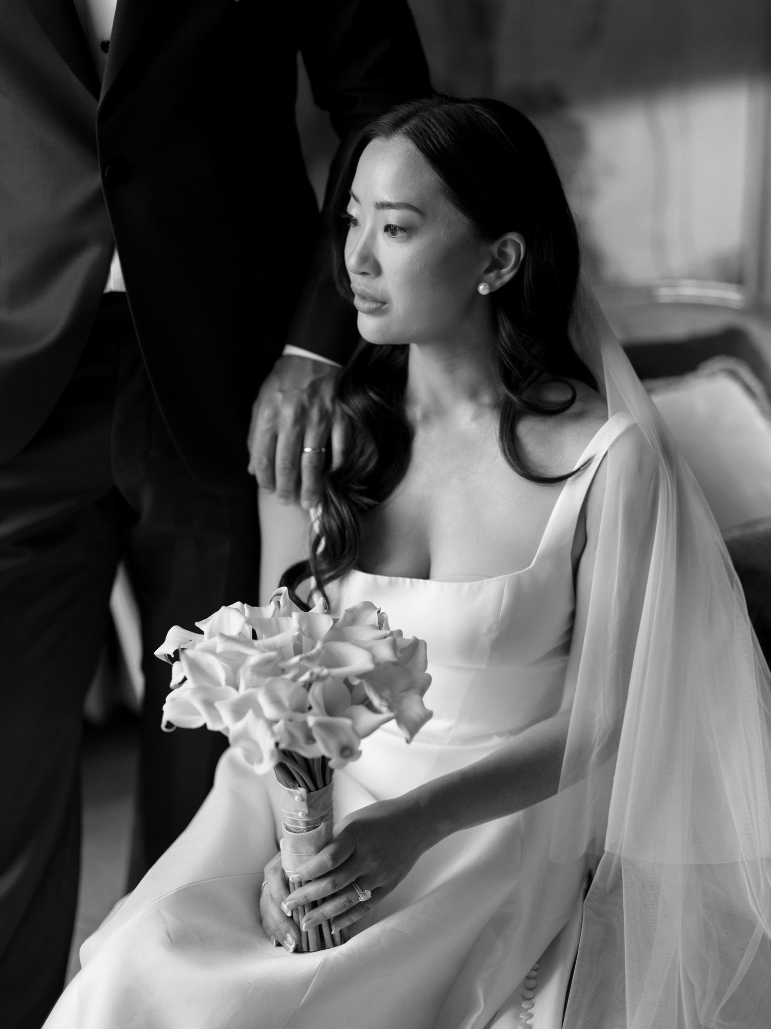 Taiwanese bride seated, holding bouquet, sunlight from window, groom's hand on shoulder.