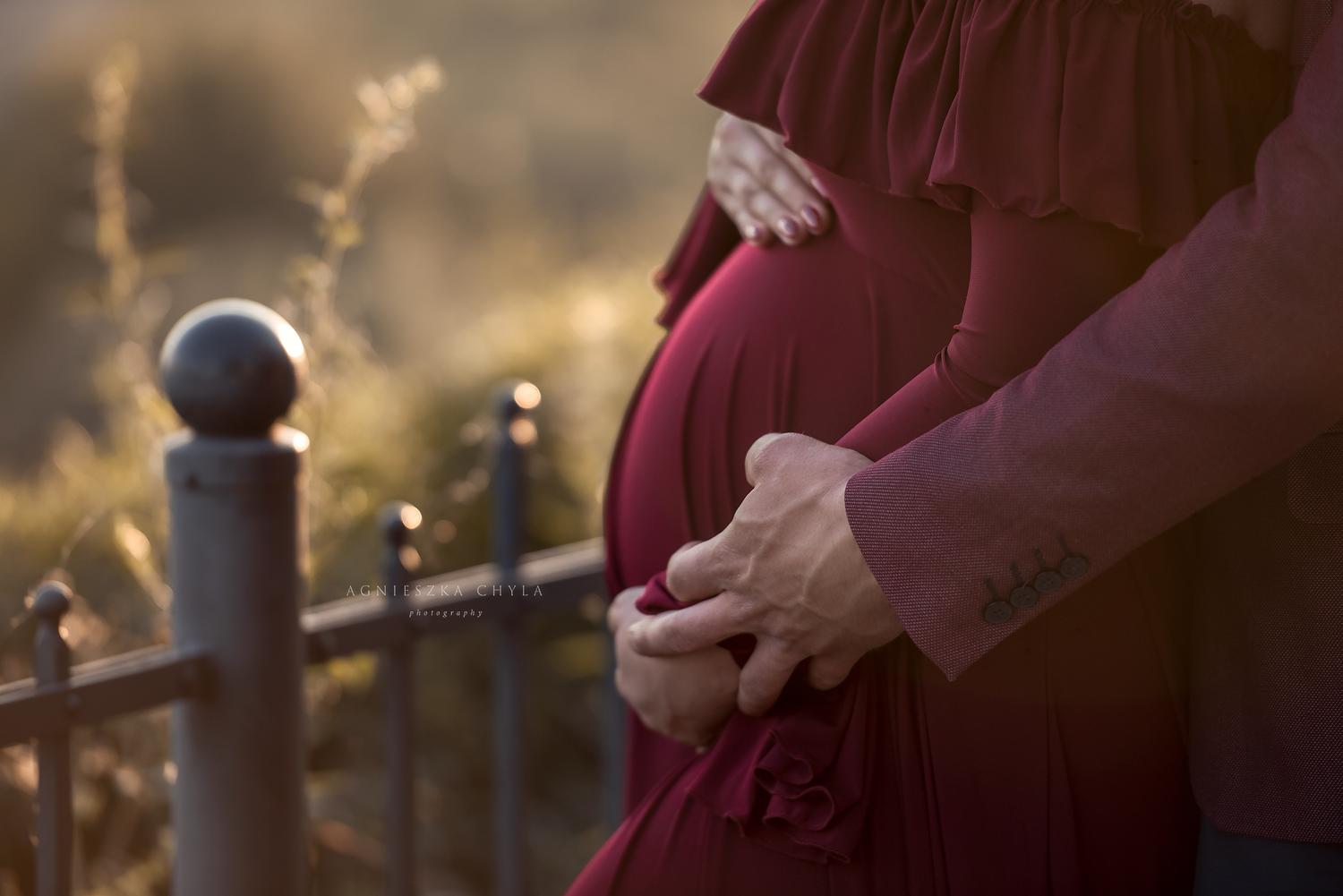 Pregnant woman in red dress cradling baby bump with partner embracing her outdoors in golden sunlight.