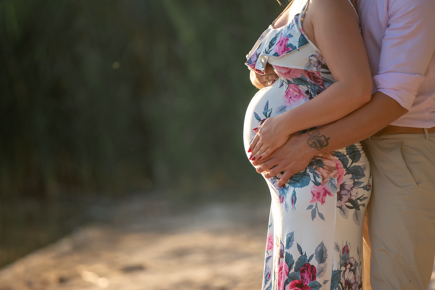 Pregnant woman in floral dress tenderly cradling baby bump with partner outdoors in warm sunlight.