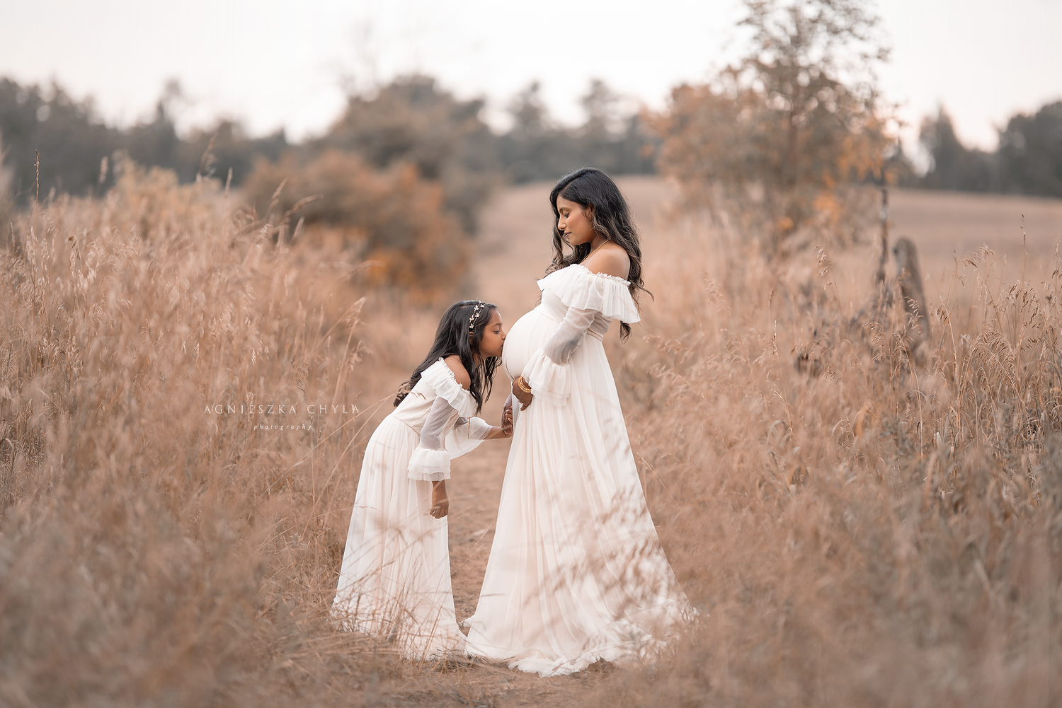 Pregnant woman in white gown with daughter in matching dress posing in golden field during maternity photo session.