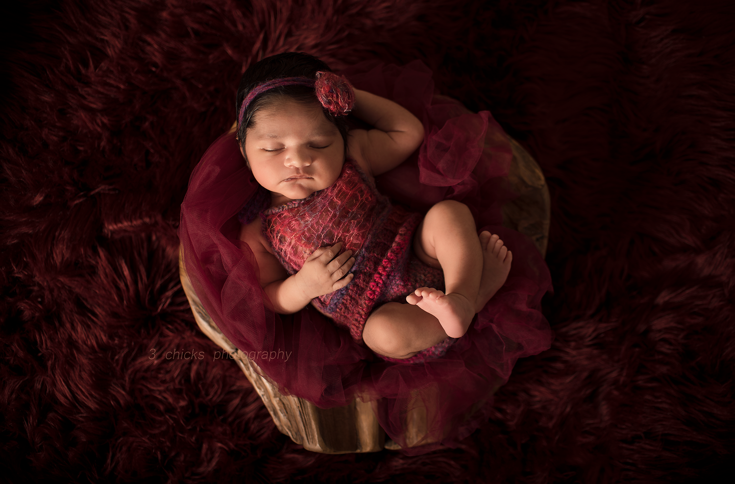 Newborn baby sleeping peacefully in a wooden bowl with burgundy fabric and fur, wearing a red knit outfit and headband.