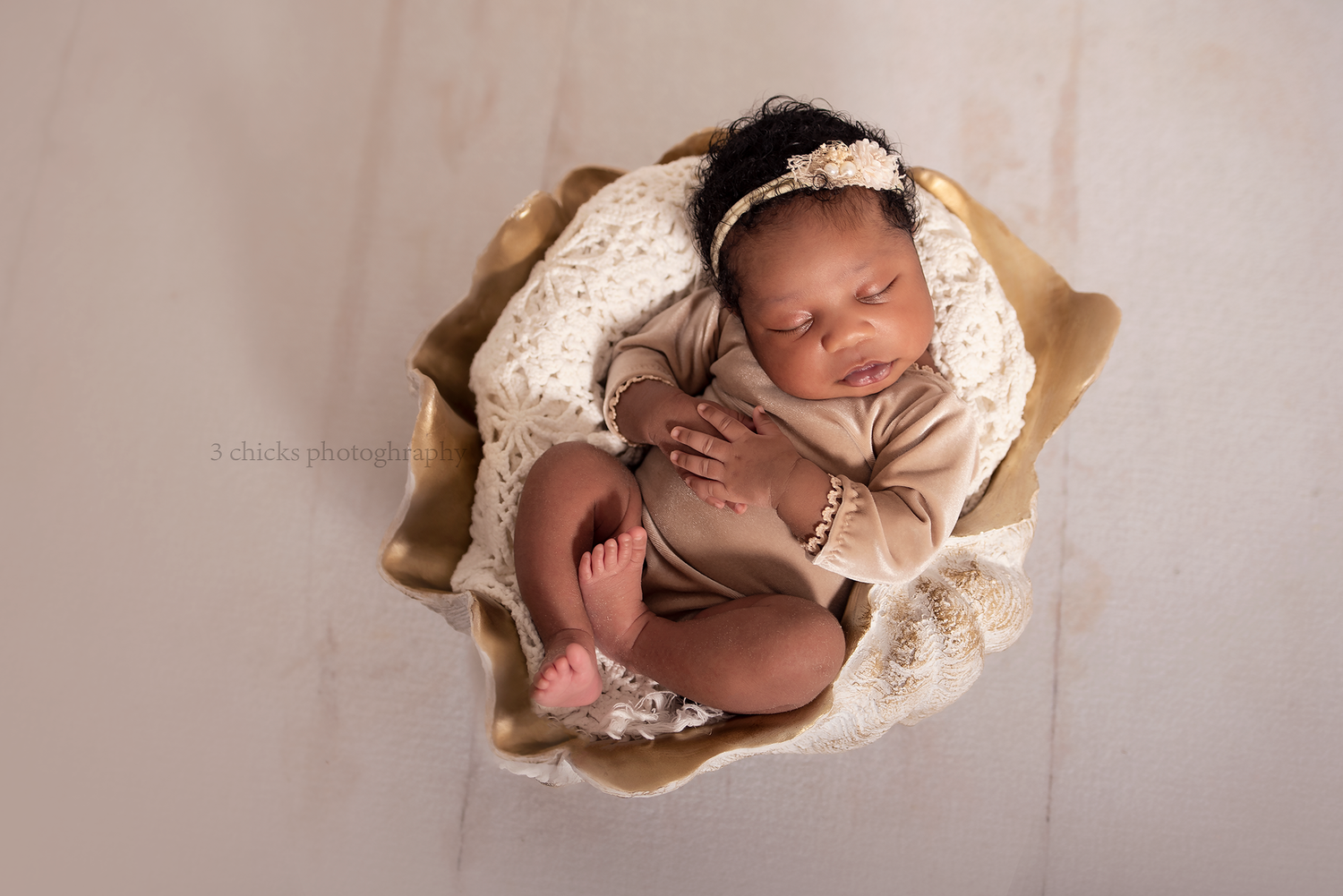 Newborn baby sleeping peacefully in a golden bowl with lace and cream wrap, wearing a floral headband.
