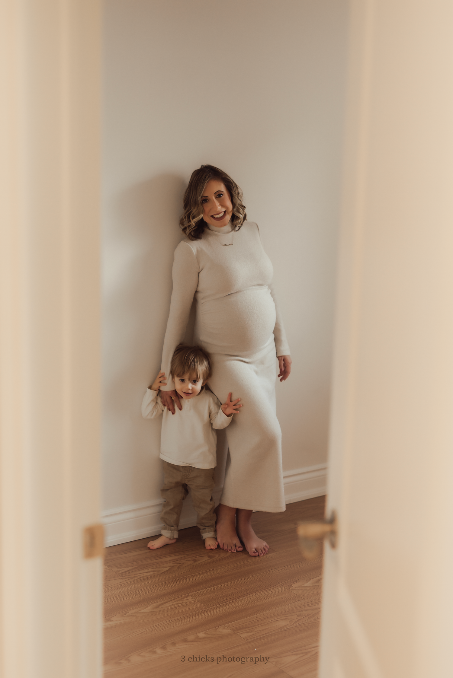 Pregnant woman in cream dress holds toddler's hands while posing in a doorway for a maternity photo session.