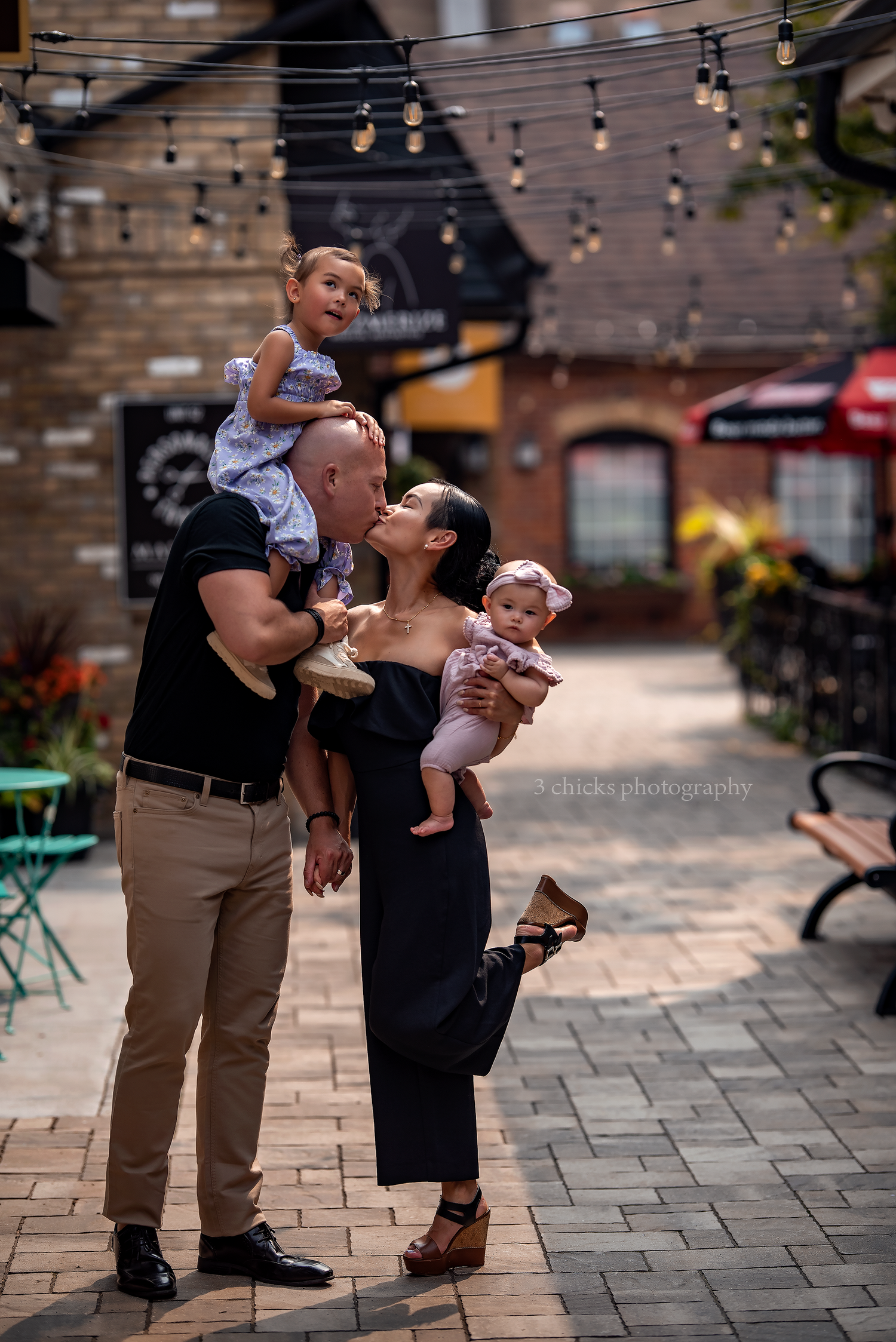 Couple sharing a kiss outdoors under string lights while holding two young children in a charming courtyard.