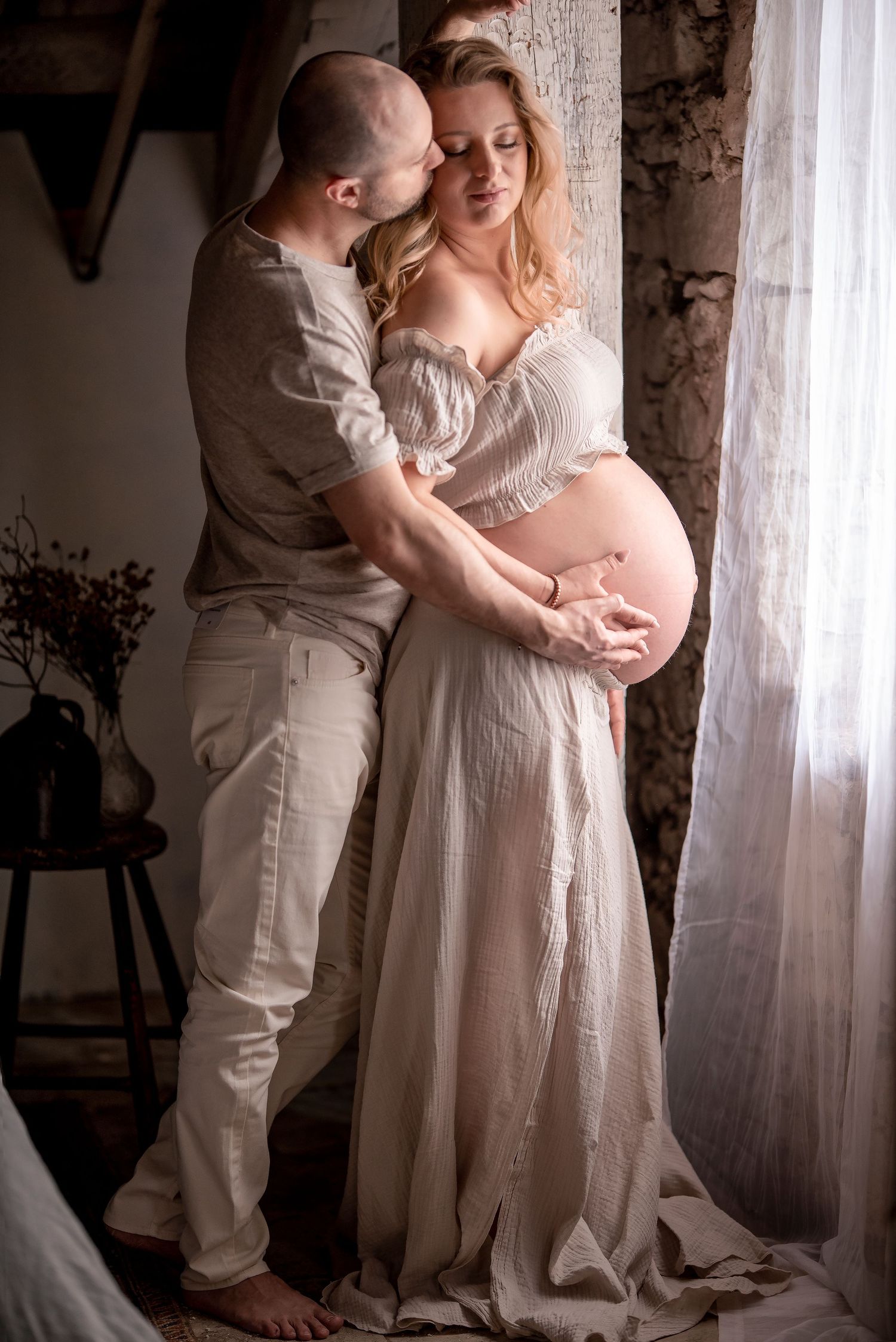 Couple sharing tender moment, man embracing pregnant woman in white dress near window in rustic room.