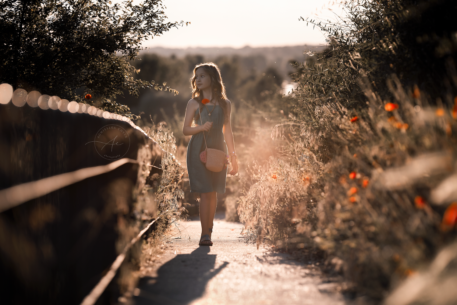 Woman walks along a sunlit path surrounded by lush greenery and wildflowers at golden hour.