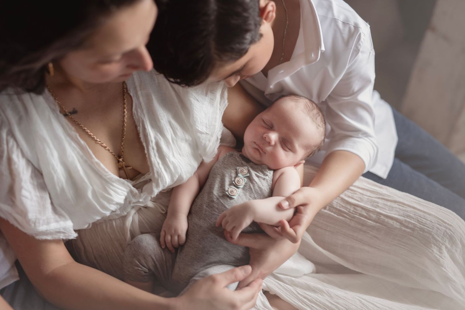 Mother in white dress kisses newborn baby while another woman holds the infant during a tender family moment.
