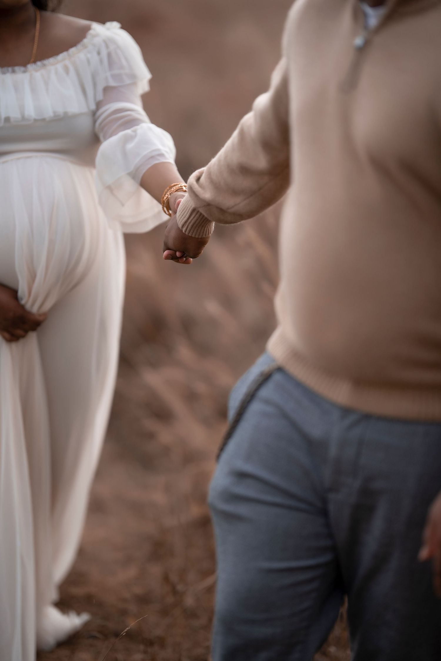 A pregnant woman in a white gown holds hands with a man in a beige sweater outdoors.