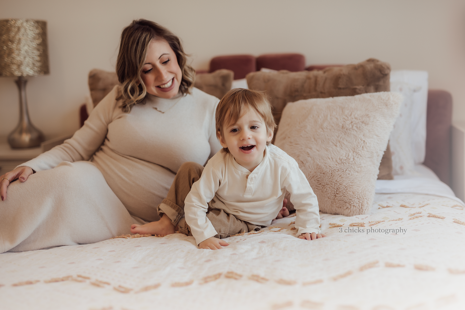 Pregnant woman in beige dress smiling on bed with laughing toddler during maternity photoshoot.