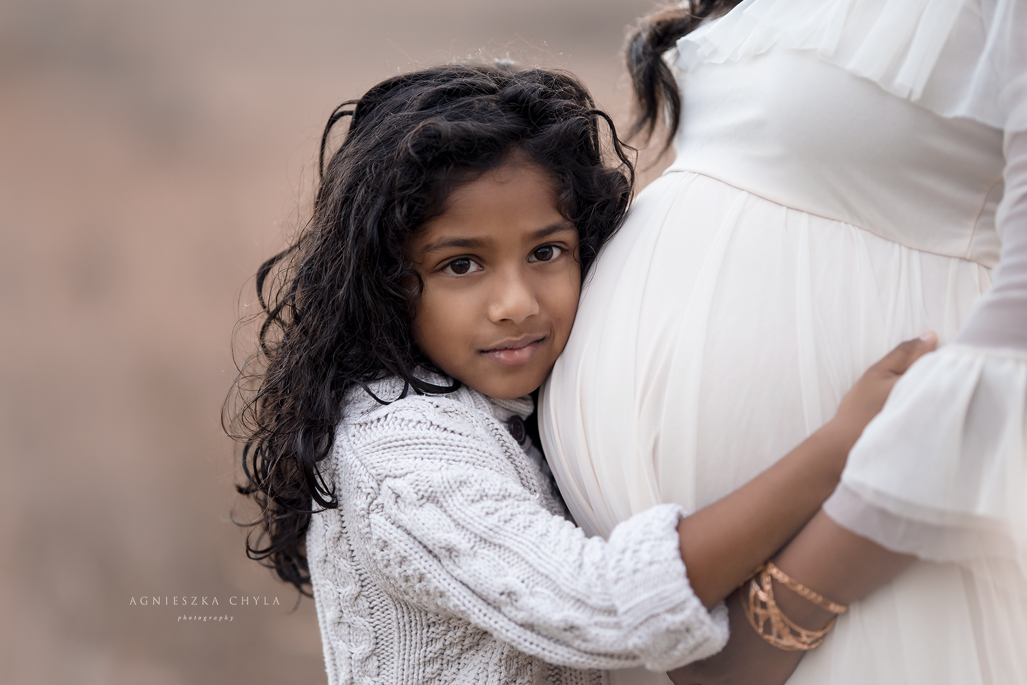 A young girl in a white sweater tenderly hugs her pregnant mother's belly during a maternity photo shoot.