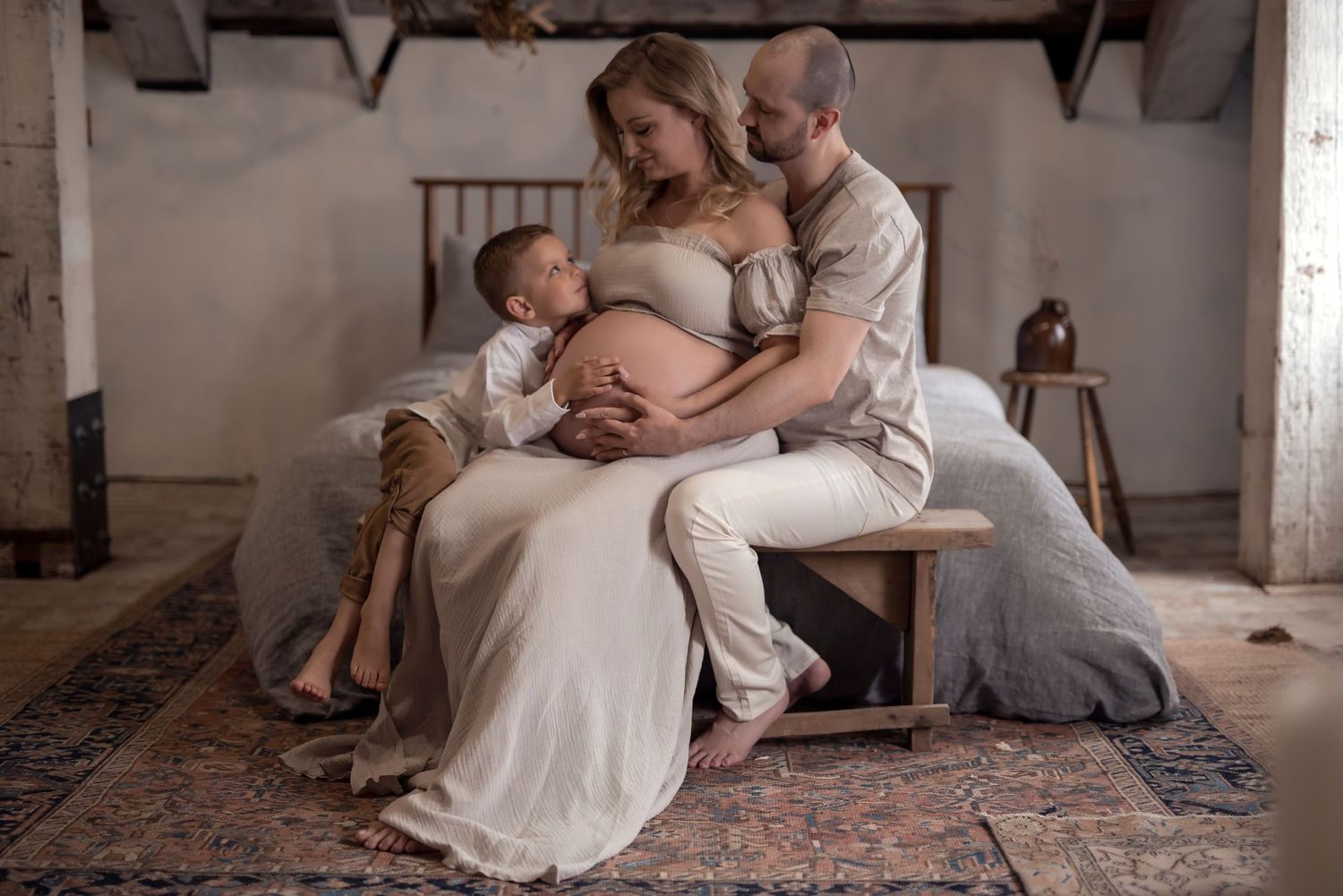 Pregnant woman in white dress sits with partner and child in rustic room during intimate maternity photo session.