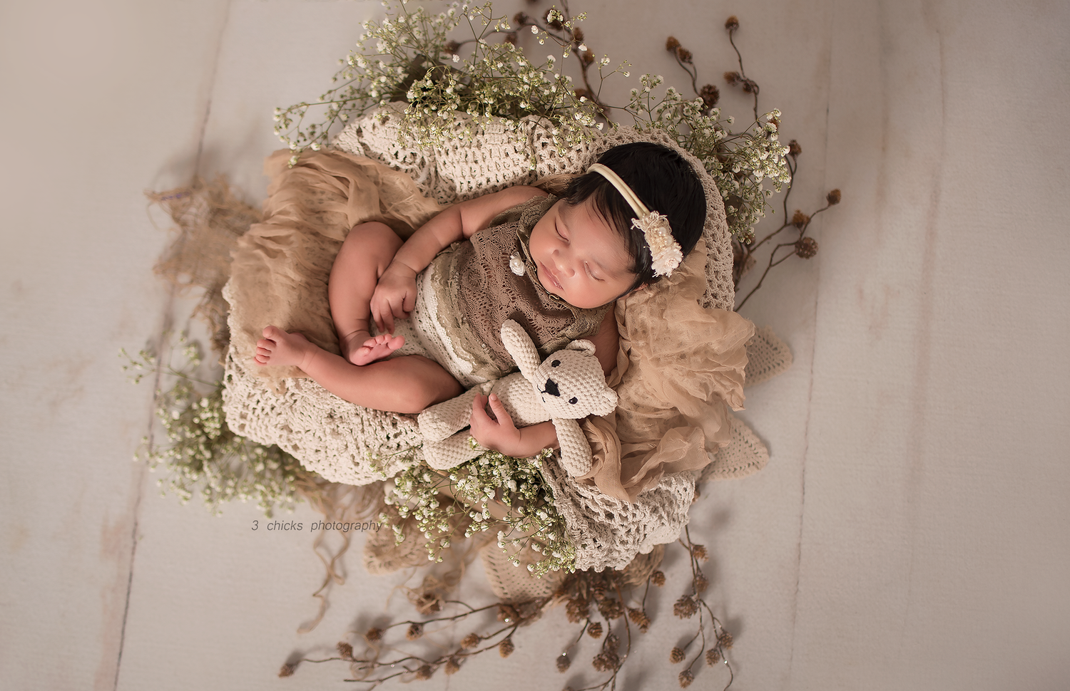 Newborn baby sleeping in a rustic woven basket with dried flowers and a tiny teddy bear, wrapped in a beige lace outfit.