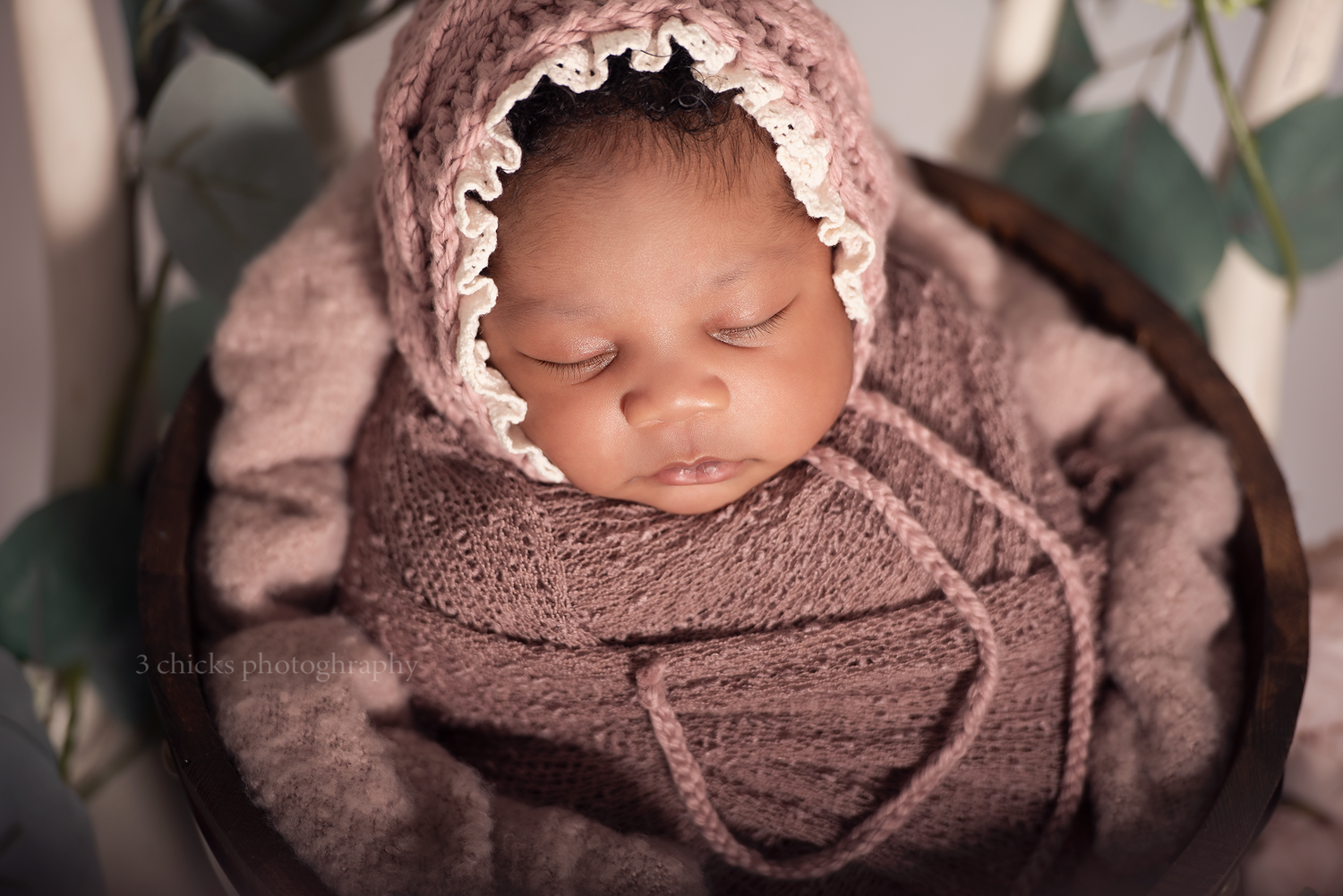Sleeping newborn swaddled in mauve wrap with lace bonnet, resting in wooden bucket with eucalyptus.