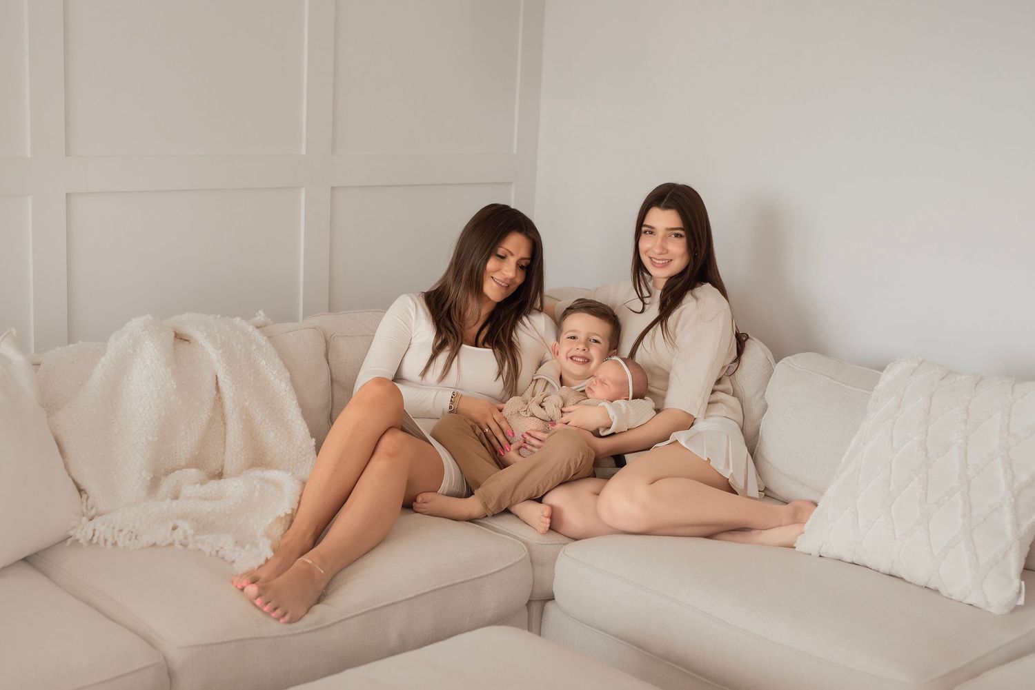 Two women and a toddler sitting together on a cream sofa in a bright white living room, wearing matching outfits.