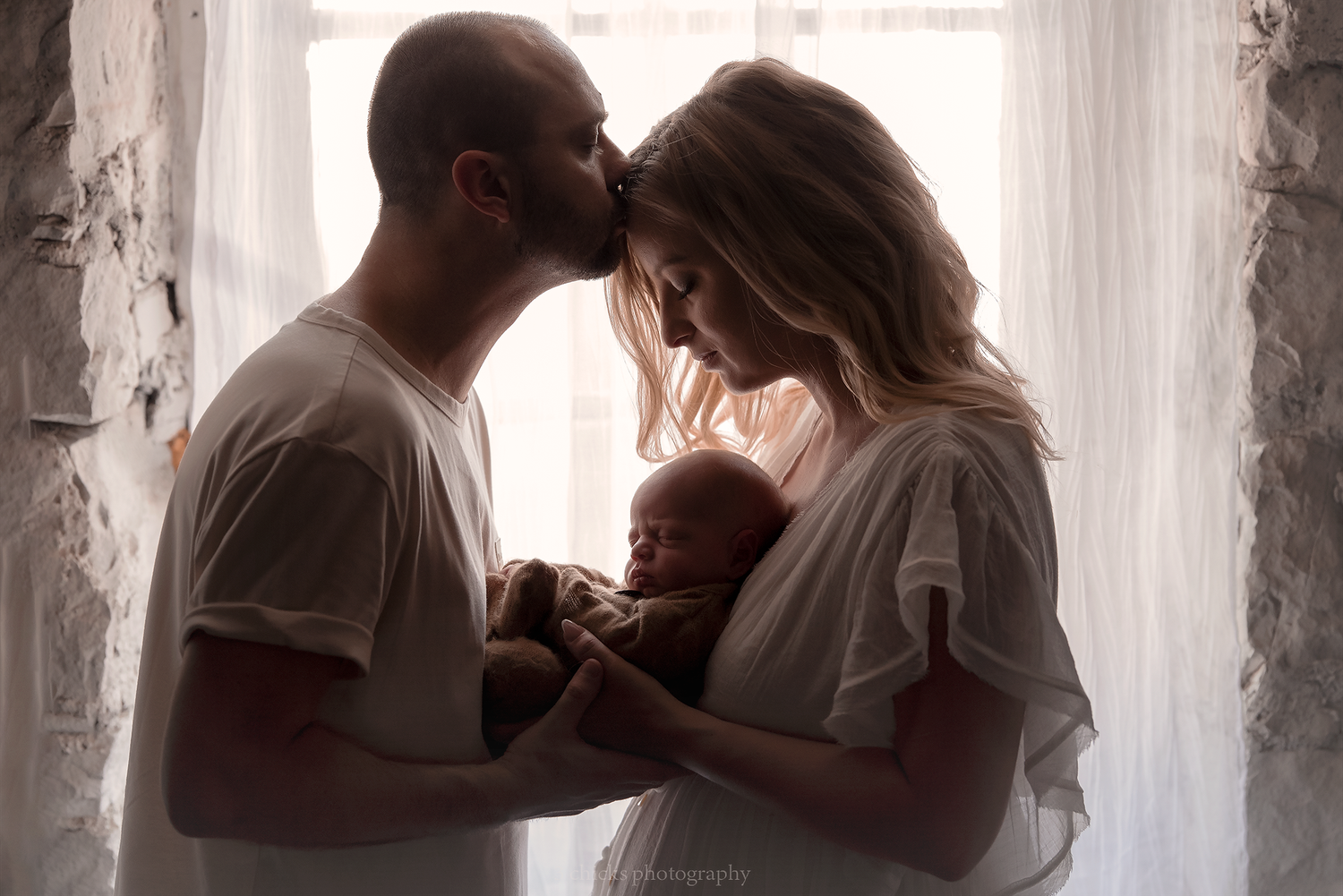 Couple sharing tender kiss while holding newborn baby in soft backlit light.