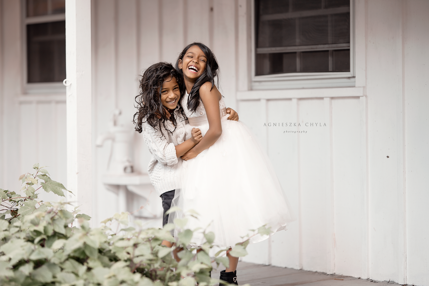 Two young girls in white dresses laugh and embrace on a white porch surrounded by greenery. Kids photography session Mississauga