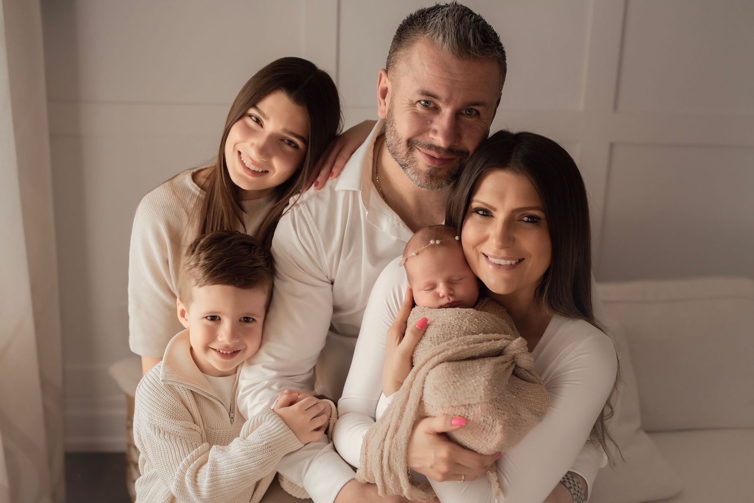 Happy family of five posing together, parents with two children and a newborn baby, wearing matching cream outfits.