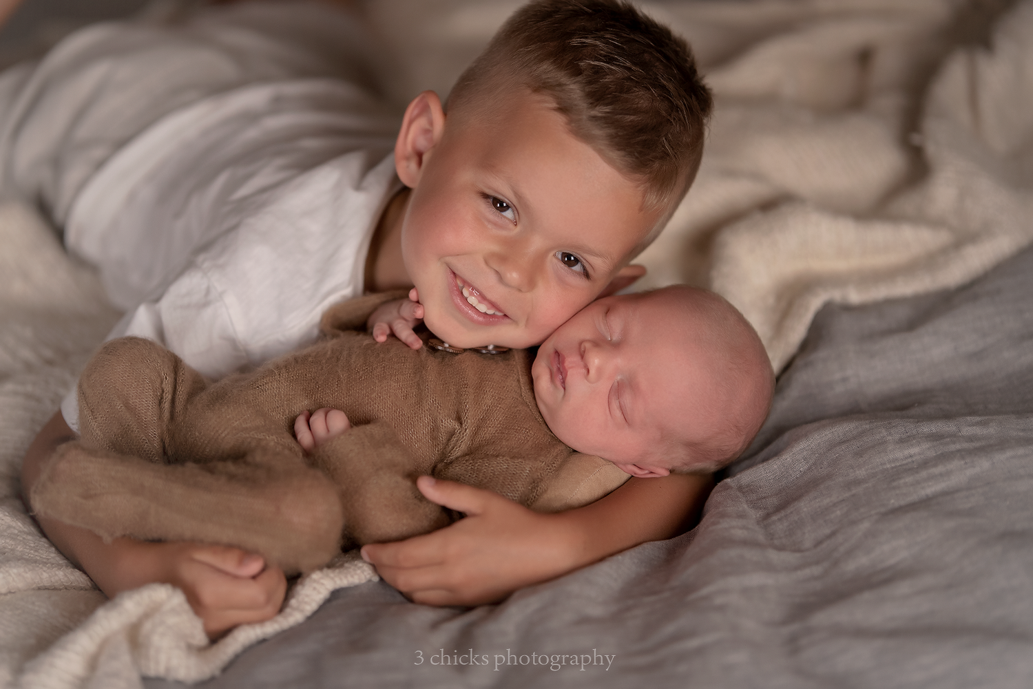 Young boy smiling while gently holding sleeping newborn baby sibling during professional photo session.